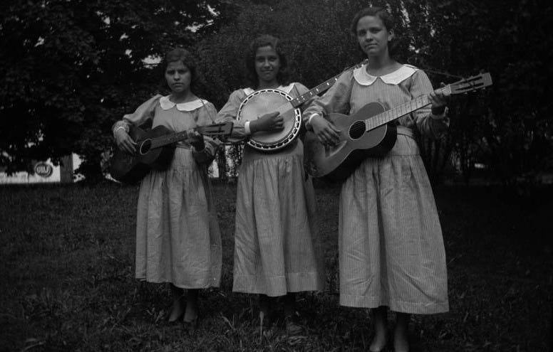 The Walters sisters, wearing matching cotton dresses with white collars, pose outdoors with musical instruments, two guitars and a banjo. 1940s.