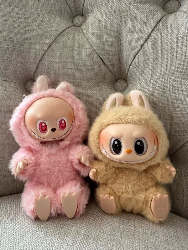Two toy dolls sit next to each other on a chair. One has pink fur and pink eyes, with its bald head sticking out of its costume, while the other is light brown and has dark eyes. 