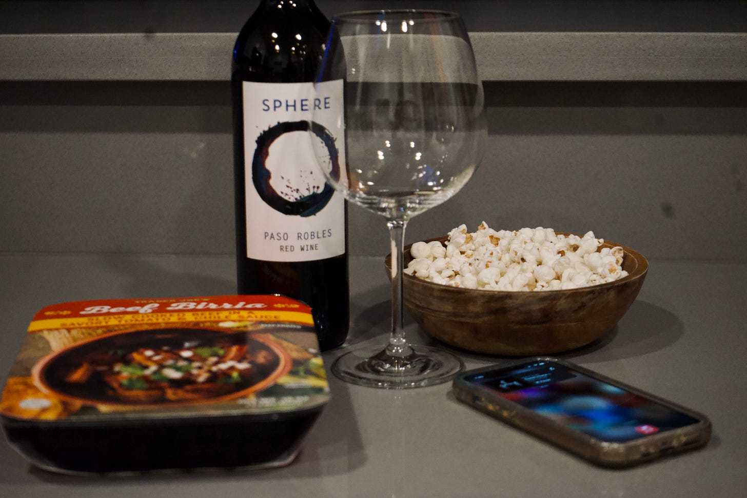 Still life on a kitchen counter featuring a bottle of red wine, an empty wine glass, a wooden bowl of popcorn, packaged dinner, and a glowing phone screen. Soft indoor lighting creates a casual evening atmosphere of solitude and unwinding.