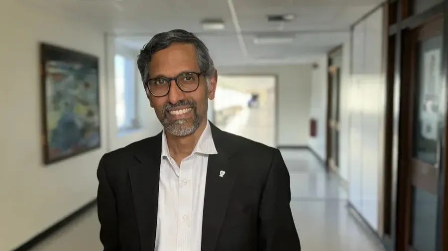 Professor Jacob George smiling, wearing a suit jacket and collared shirt in a hallway.