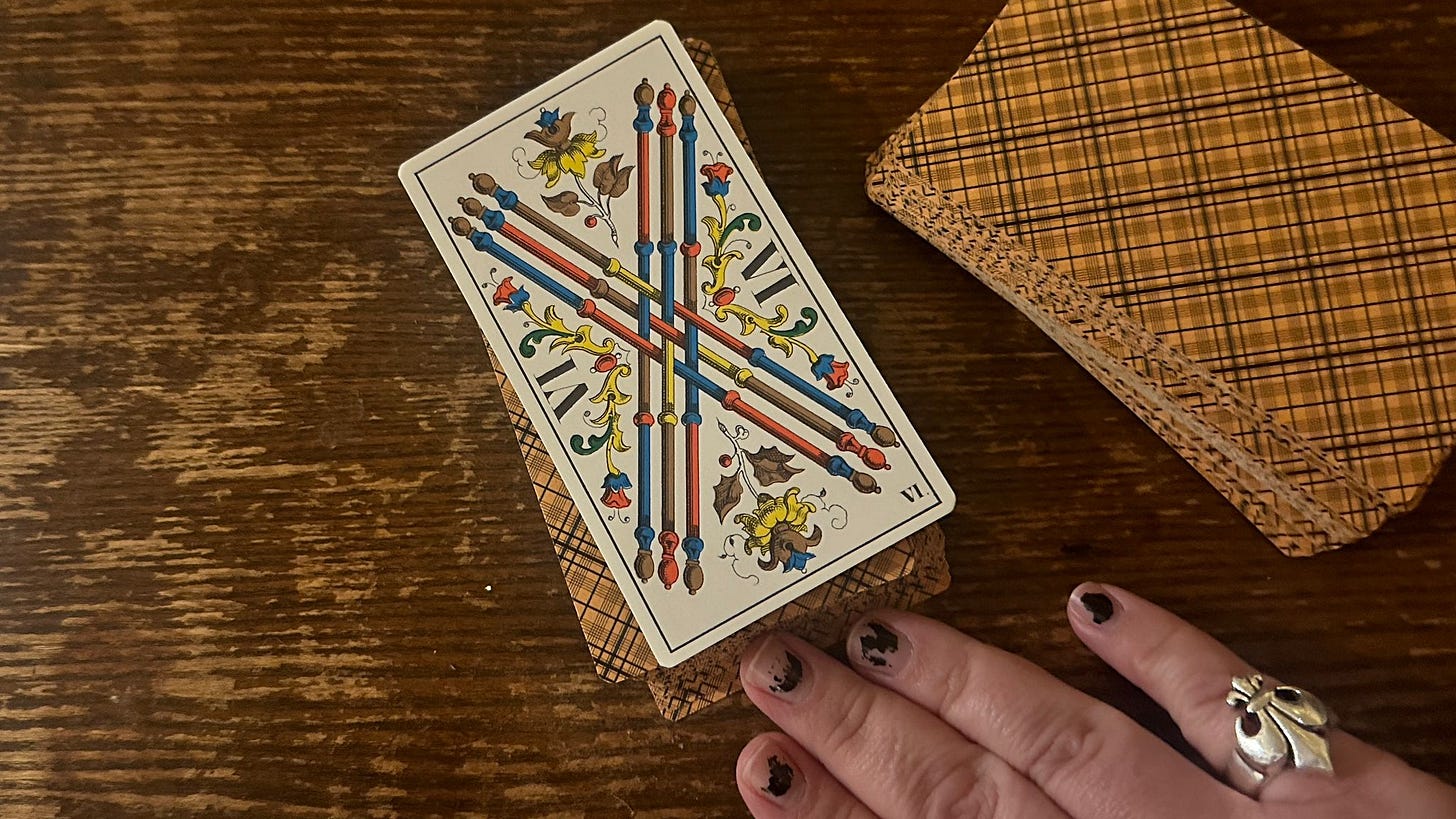 Six of Wands (VI de Bâtons) from Tarot de Marseille deck resting on patterned card backs with hand wearing rings visible, photographed for the Tarot DMs interview with Alice Slater by Finbarre Snarey.