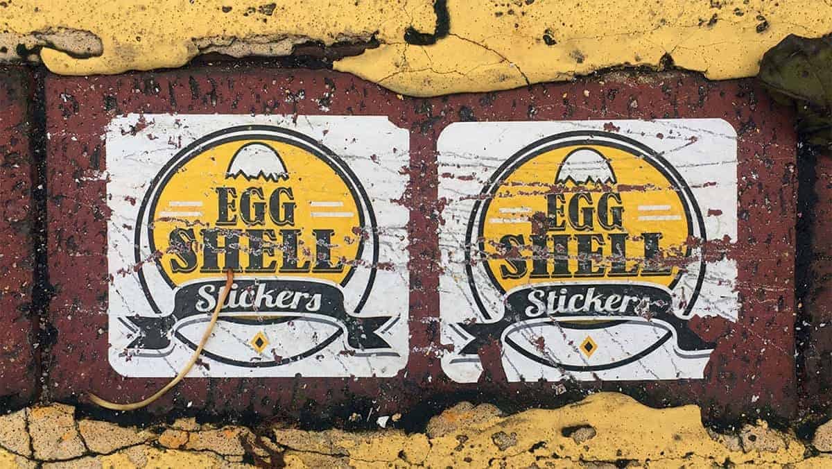 Two weathered 'Egg Shell Stickers' placed side by side on a rough, red brick surface with peeling yellow paint around the edges. The stickers feature a yellow egg logo with black and white text.