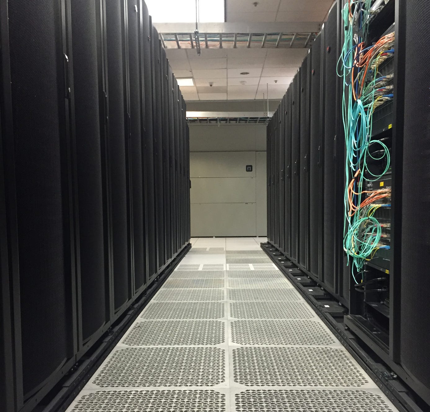 A long aisle in a data center lined with tall black server racks on both sides, with visible cables and a raised floor.