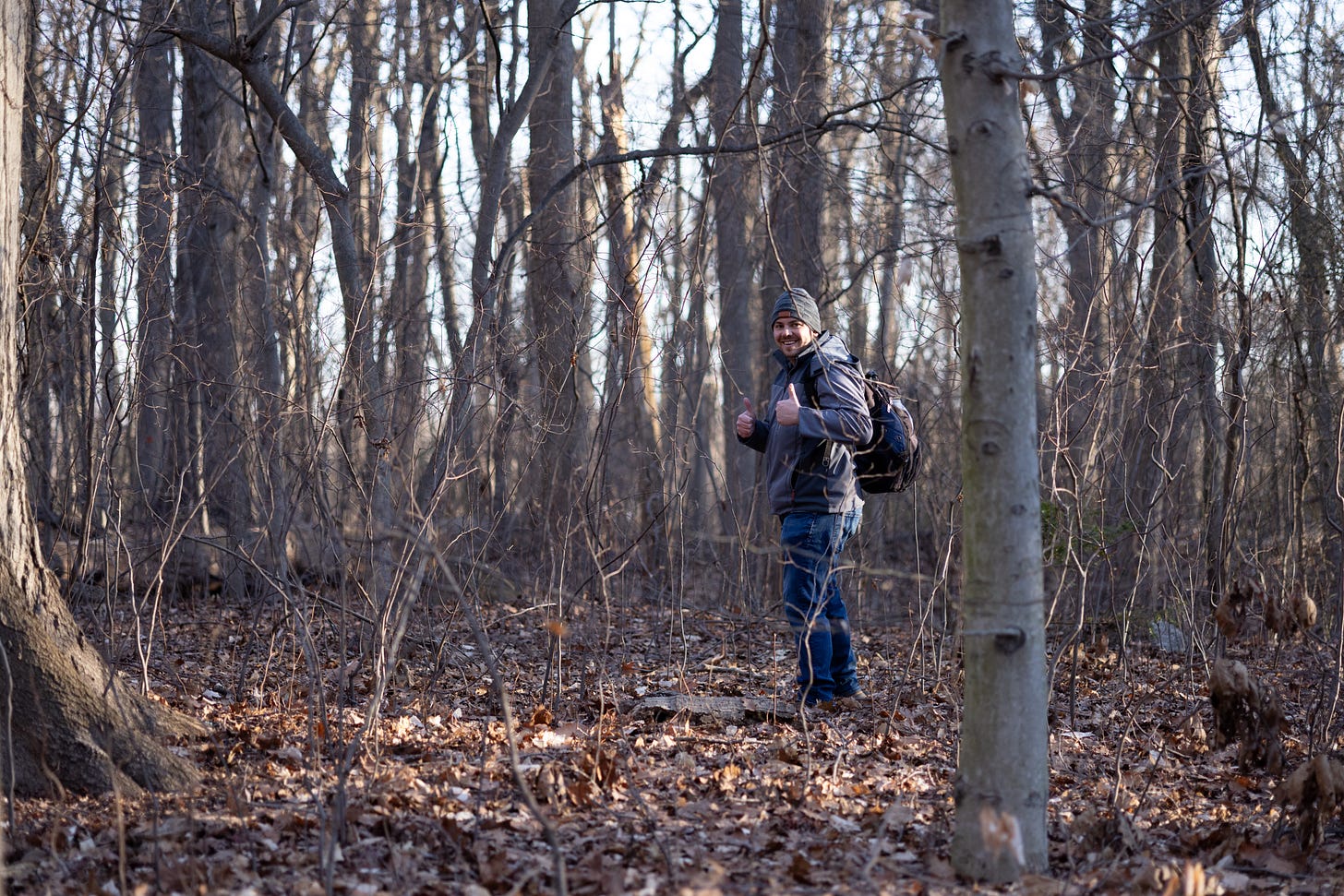 a forested scene with lots of leaf litter and a man at middle distance in early spring weather wear looking back at the camera giving a thumbs up and smiling a forested scene with lots of leaf litter and a man at middle distance in early spring weather wear looking back at the camera giving a thumbs up and smiling