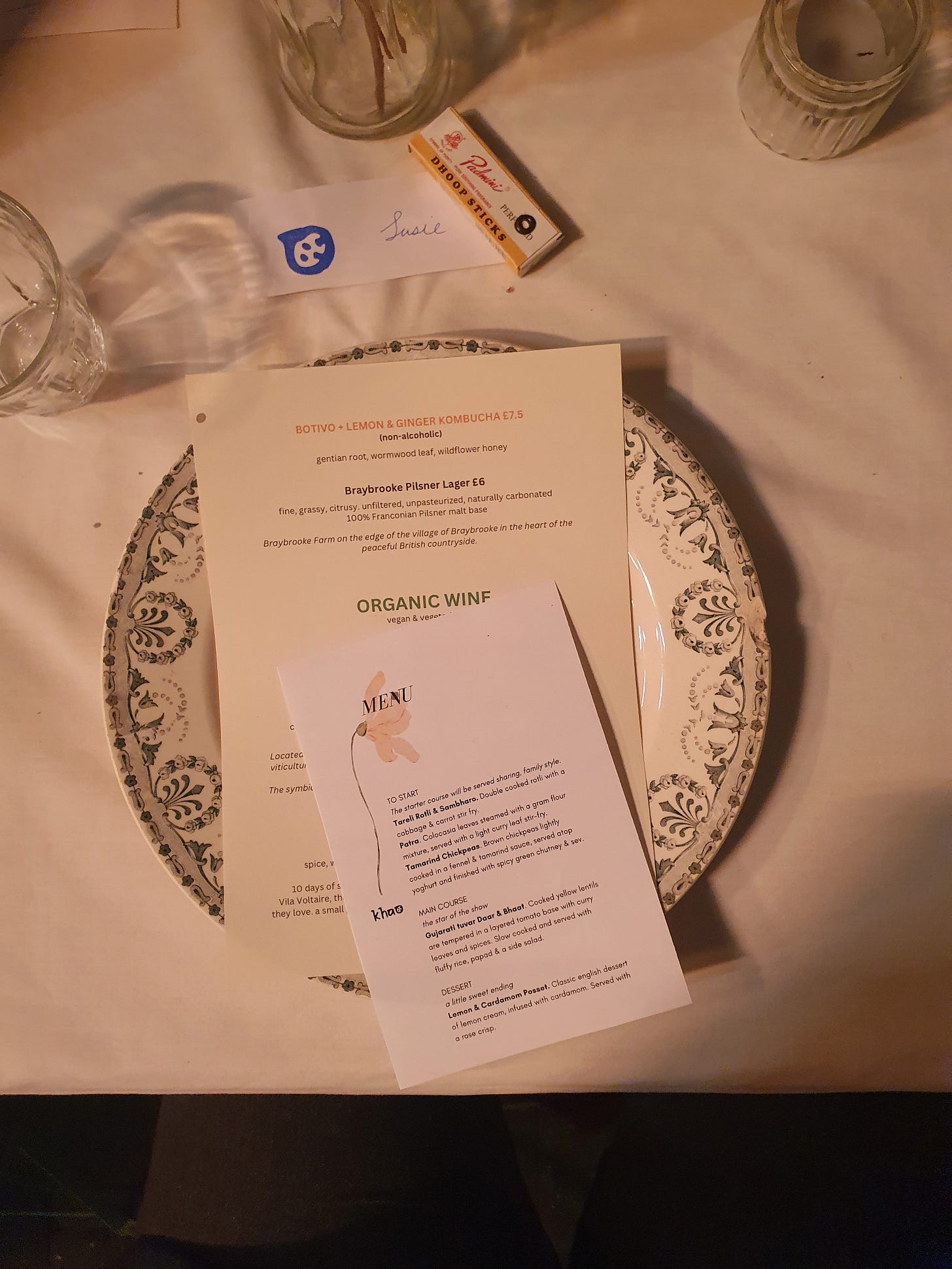 A plate on a table spread with a white tablecloth. On top of the plate is a drinks menu and the menu for the evening.