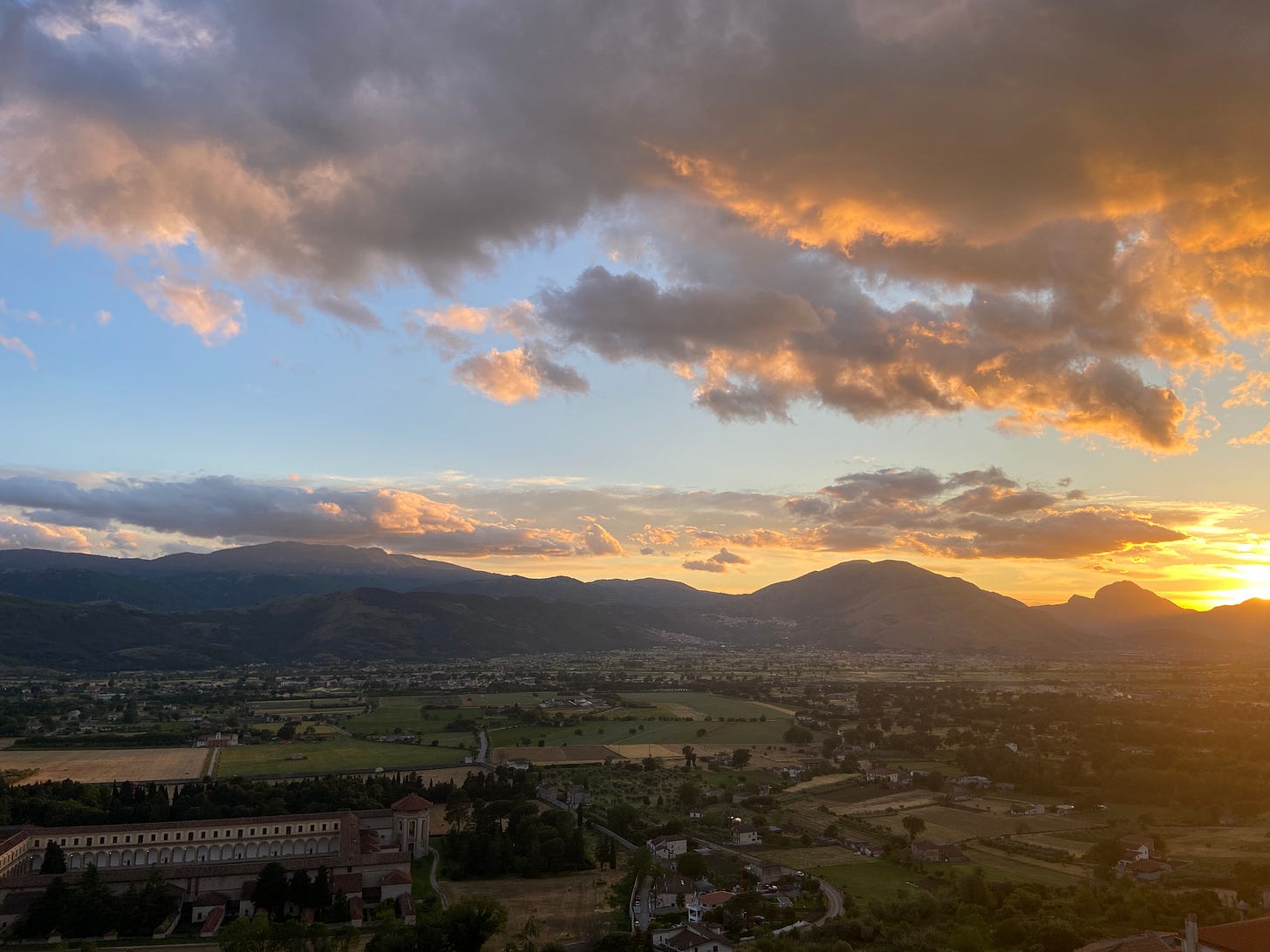 A setting sun behind a mountain, a valley below filled with fields and buildings. 