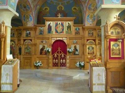 Interior of a wood-paneled church with a carved wooden iconostasis, rows of icons, and hanging oil lamps, doors open toward the altar.