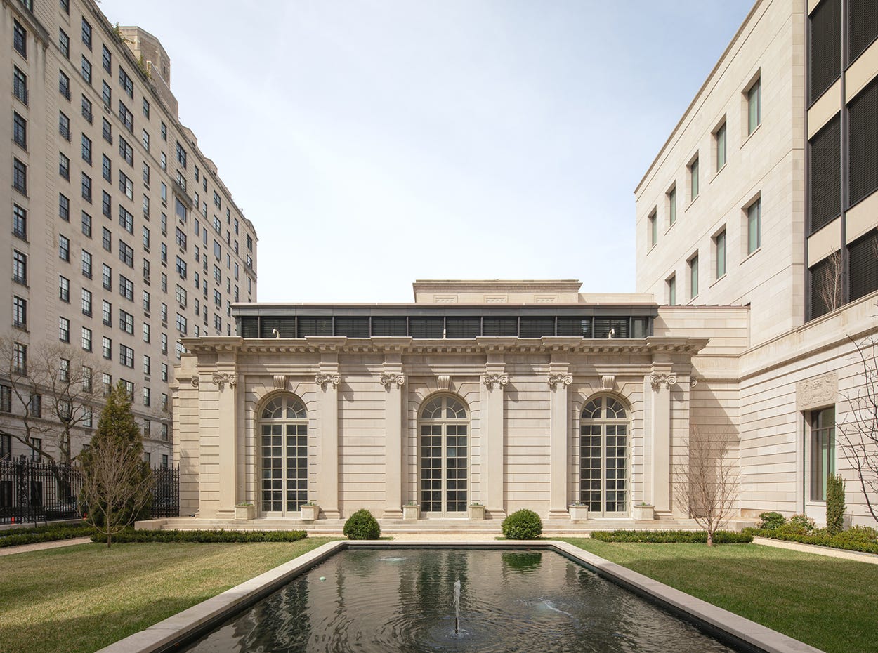 A regal yet austere neoclassical facade glows as it looks over a manicured lawn and reflecting pool, flanked by two taller buildings in similar sandy shades.
