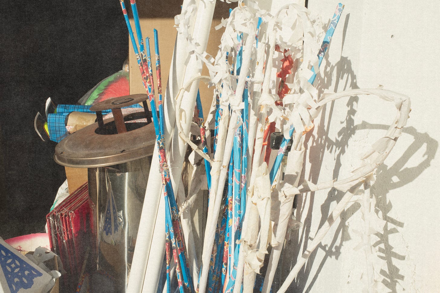 Colour photograph showing funeral offerings displayed outside a shop. White paper stakes cast distinct shadows on a white wall. The scene is brightly lit, bordering on overexposed. In the background, paper legs—part of a human figure—are visible.
