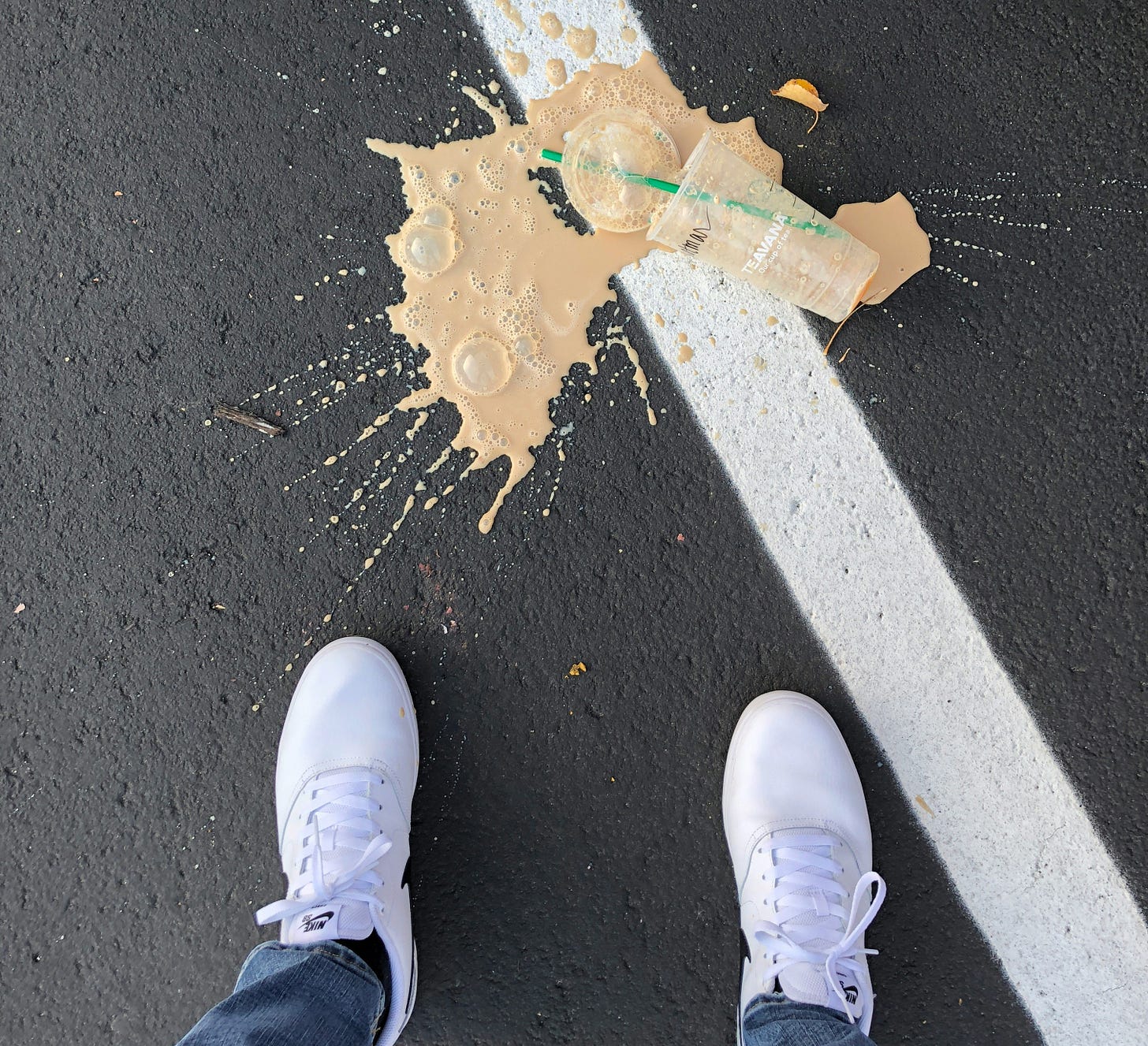 Spilled coffee on asphalt next to white sneakers and a white line. Transparent cup with a green straw lies on the ground, creating a messy scene.