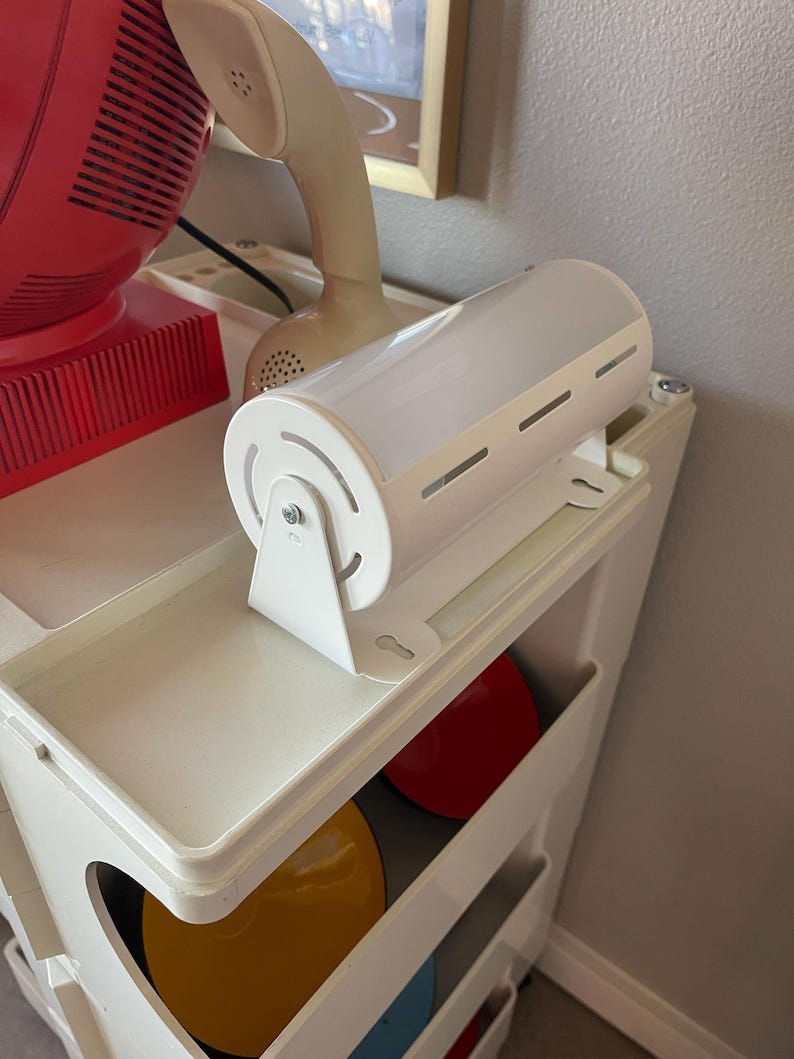 May include: Close-up view of a white cylindrical LED light fixture mounted on a white plastic shelving unit. The light has a frosted white cover and several small ventilation slots. The shelving unit holds colorful round objects, possibly records or discs, in red, yellow, and light blue.  A vintage-style off-white telephone and a red electronic device are visible in the background. May include: Close-up view of a white cylindrical LED light fixture mounted on a white plastic shelving unit. The light has a frosted white cover and several small ventilation slots. The shelving unit holds colorful round objects, possibly records or discs, in red, yellow, and light blue.  A vintage-style off-white telephone and a red electronic device are visible in the background.