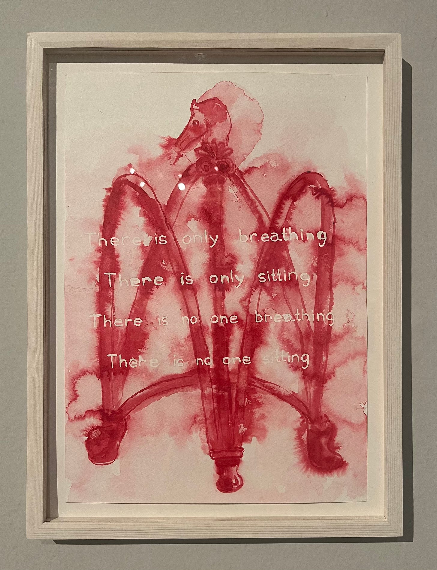 A framed print in red ink is on a greyish wall. The print depicts an abstract piece of furniture with a horse's head and is overlaid with hand-painted words: "There is only breathing / There is only sitting / There is no one breathing / There is no one sitting"