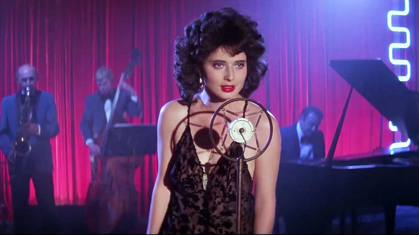 Still from the film Blue Velvet (1986). A woman with dark, voluminous hair and red lipstick stands at a vintage microphone, wearing a black lace dress. Behind her, a jazz band performs on a dimly lit stage with deep red curtains and moody blue lighting. The setting exudes a dreamlike, noir atmosphere, capturing the film's surreal and mysterious tone.