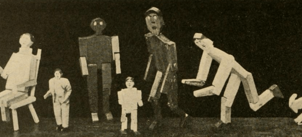 Black and white photograph of figures made out of what appears to be cardboard. Four larger figures appear to be looking at two smaller ones.