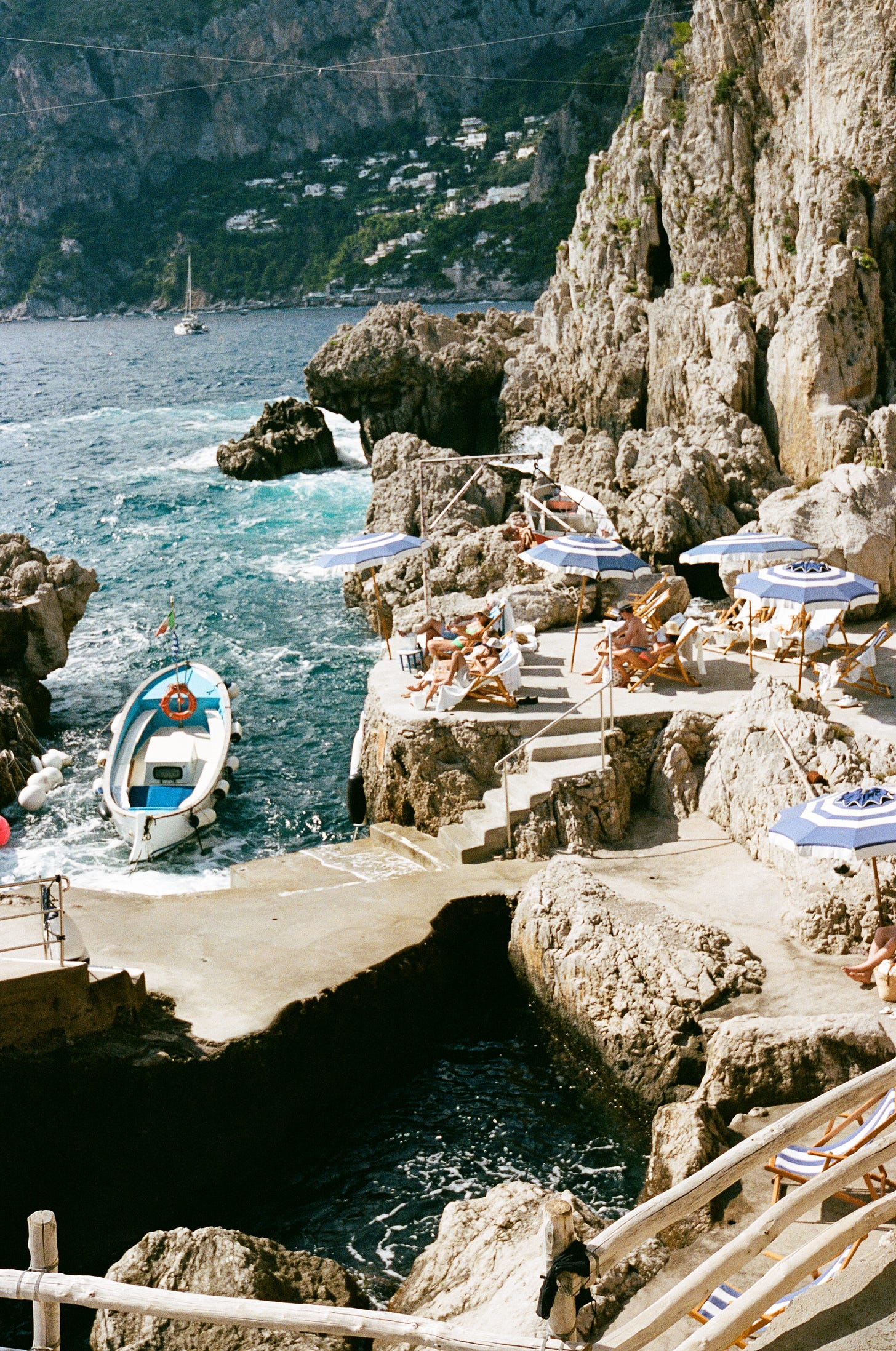 An image of the coast in Italy with boats, and umbrellas and chairs on rocks.