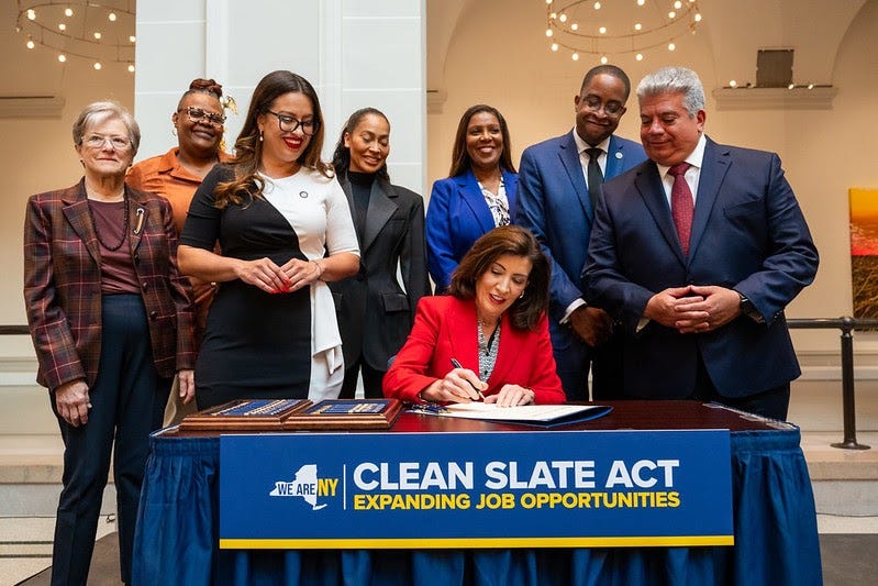 Governor Kathy Hochul sits at the Clean Slate signing table, surrounded by legislators and supporters. Governor Kathy Hochul sits at the Clean Slate signing table, surrounded by legislators and supporters.