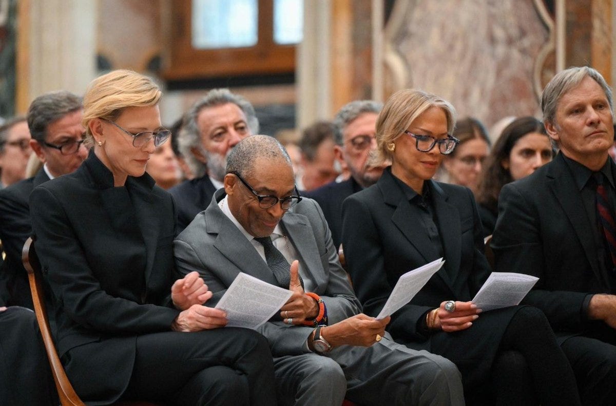 Four individuals seated in a row in a grand hall with ornate walls and columns. Cate Blanchett on the left wearing glasses and black attire holding papers. Spike Lee next to her in gray suit gesturing with hand wearing a watch and bracelet holding papers. Another woman in black with glasses holding papers. Viggo Mortensen on the right in black suit with red tie holding papers. Surrounding audience members in formal wear seated on chairs. Four individuals seated in a row in a grand hall with ornate walls and columns. Cate Blanchett on the left wearing glasses and black attire holding papers. Spike Lee next to her in gray suit gesturing with hand wearing a watch and bracelet holding papers. Another woman in black with glasses holding papers. Viggo Mortensen on the right in black suit with red tie holding papers. Surrounding audience members in formal wear seated on chairs.