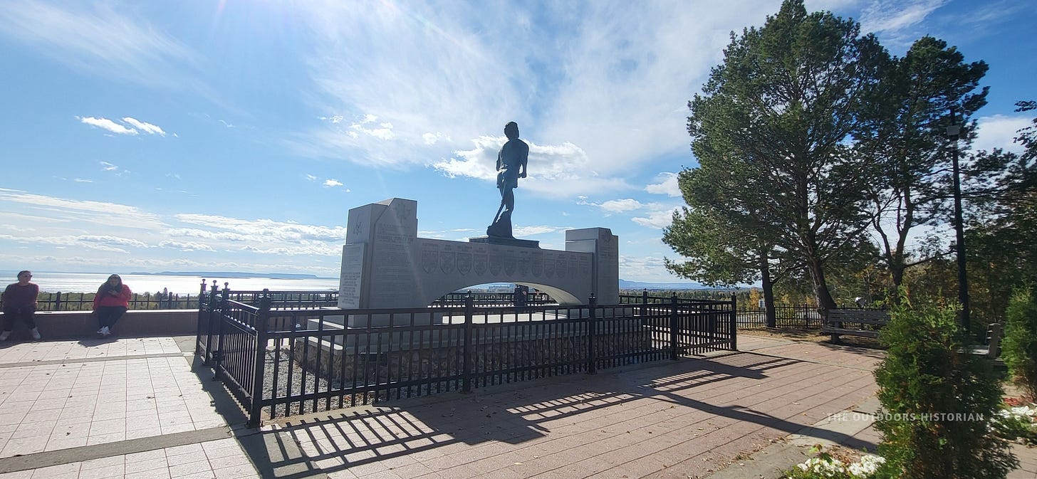 A bronze statue of Terry Fox mid-stride stands on a granite pedestal. In the background, the silhouette of the Sleeping Giant formation is visible across the blue water of Lake Superior.