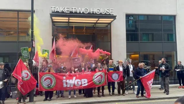 IWGB members holding a red banner emblazoned with the union's logo are shown picketing outside Take-Two House, the company's London HQ. People are shown holding signs, flags and megaphones as red and yellow smoke rises from smoke grenades.
