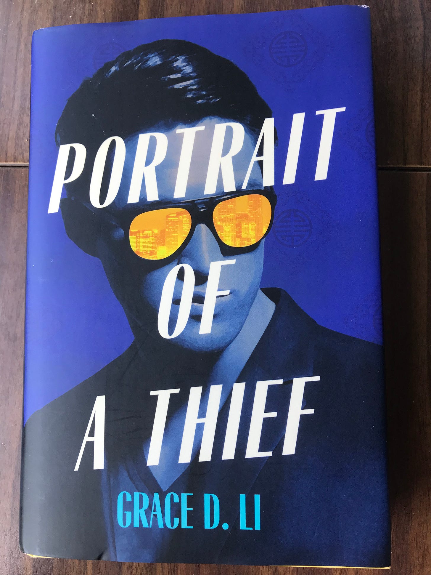 A book on a wooden table. The book cover is electric blue, with the title "Portrait of a Thief" in white capital letters slanted slightly upward to the right, superimposed on an image of a man in a suit jacket and tshirt, and black hair slicked back wearing sunglasses with bright yellow lenses with city buildings in the reflection.