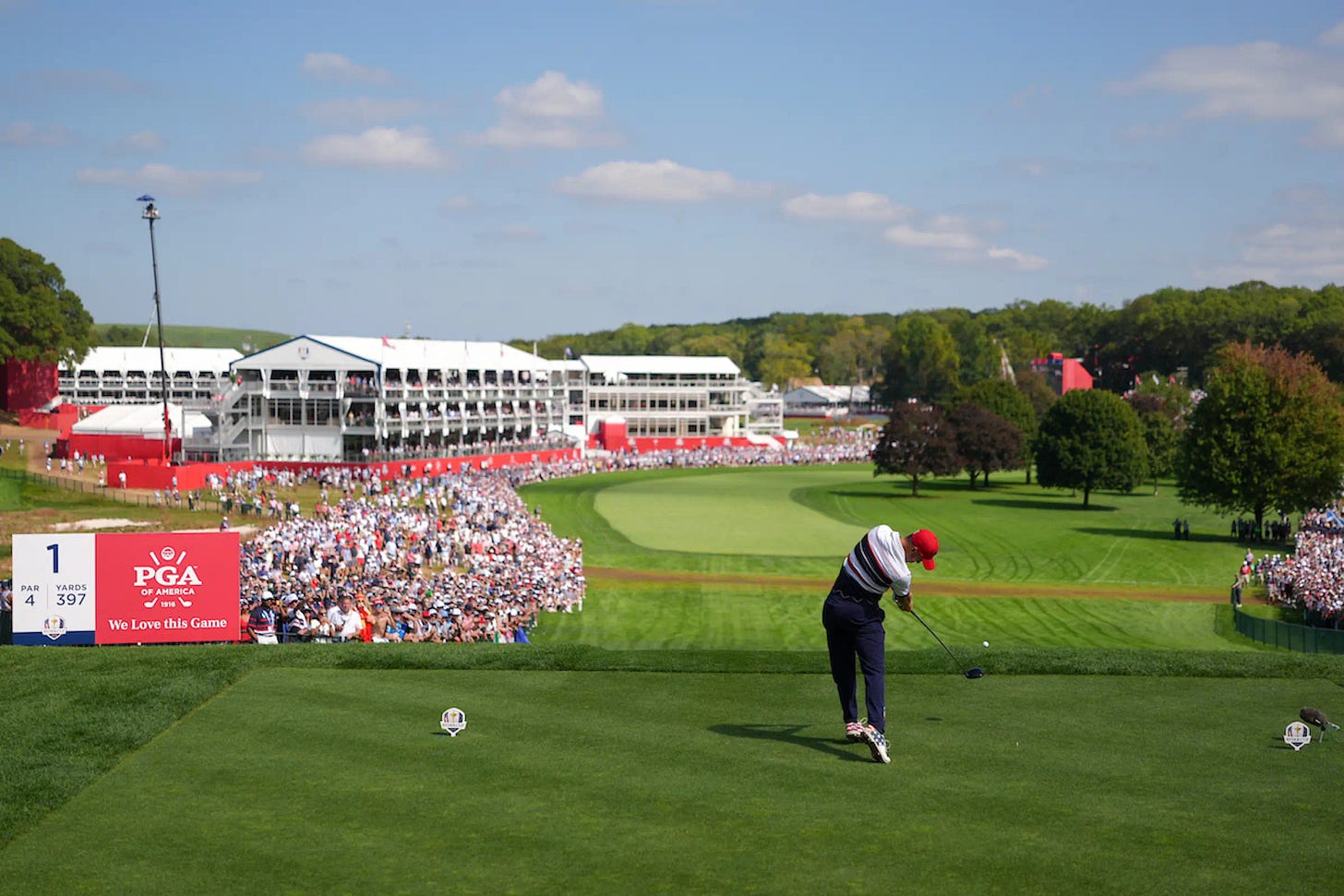 A Team USA golfer tees off on the opening hole of the 2025 Ryder Cup at Bethpage Black, with a packed crowd and grandstands lining the course.