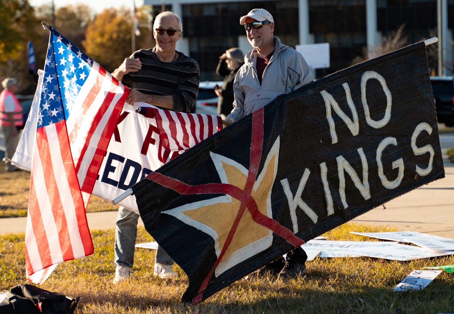 Two older men stand on a grassy roadside at a daytime protest, smiling as they hold American flags and a large black banner painted with a gold star, red X, and the words “NO KINGS,” with other demonstrators and signs blurred in the background. Two older men stand on a grassy roadside at a daytime protest, smiling as they hold American flags and a large black banner painted with a gold star, red X, and the words “NO KINGS,” with other demonstrators and signs blurred in the background.
