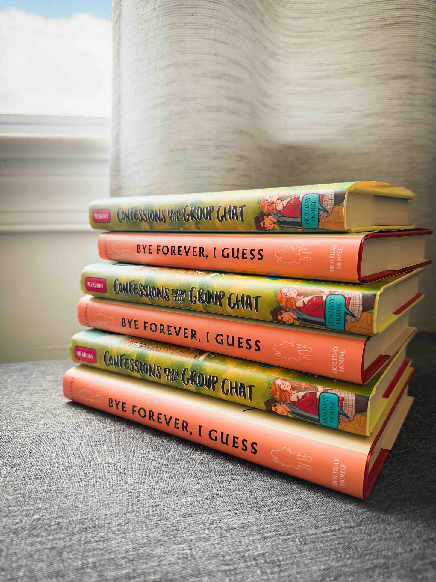 A stack of books: bye forever, i guess and confessions from the group chat. The books alternate. They're sitting on a gray surface, and a window in the background has a cloud. 
