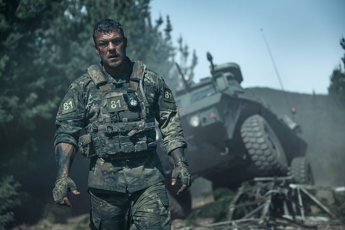 Alan Ritchson as the soldier 81, standing tired in front of a crashed armored vehicle. 