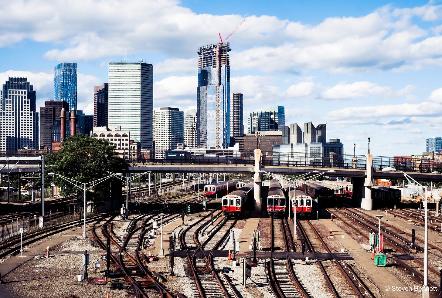 olor photo of the Boston MBTA rail yard and skyline from the West 4th Street bridge, with red-and-white subway cars and clouds overhead. olor photo of the Boston MBTA rail yard and skyline from the West 4th Street bridge, with red-and-white subway cars and clouds overhead.