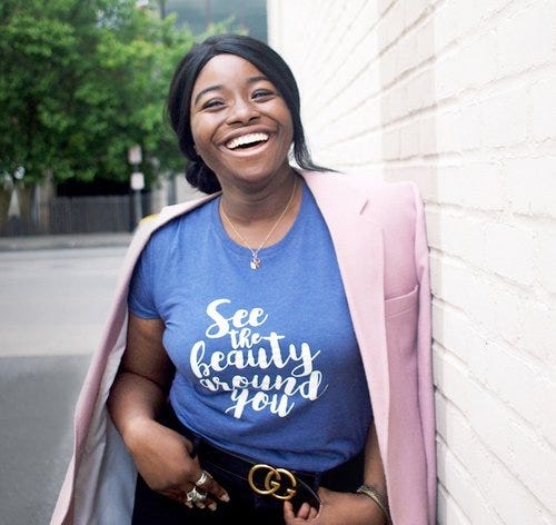 woman with black hair, pink jacket and blue shirt leans against white wall smiling with green trees in background