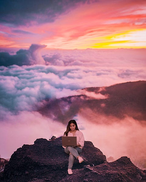 woman sits with her laptop on top of a mountain, above the clouds, at sunrise.