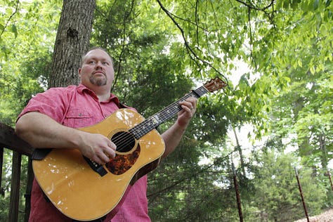 Eric Beaty playing guitar posing with Breedlove and Ibanez guitars wearing a NaNoWriMo winner shirt and posing with Smart Focus book series