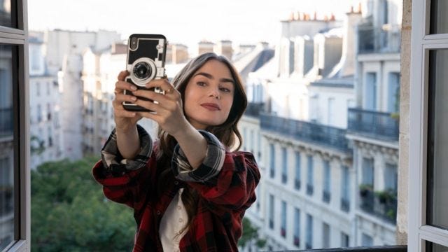 Lily Collins, interpretando Emily, tira selfie de sacada em Paris, com prédios históricos ao fundo