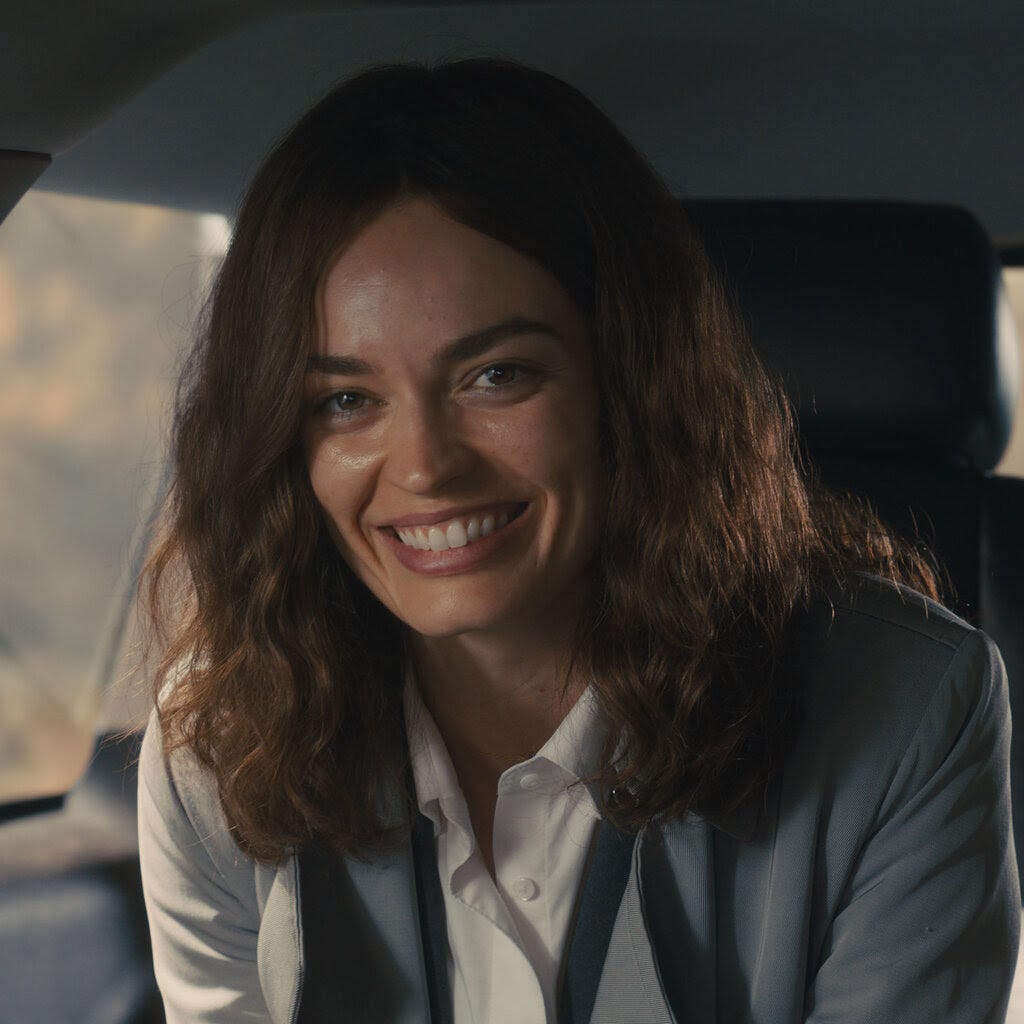 A woman with wavy brown hair and a light gray blazer over a white shirt smiles while sitting in the back seat of a car.