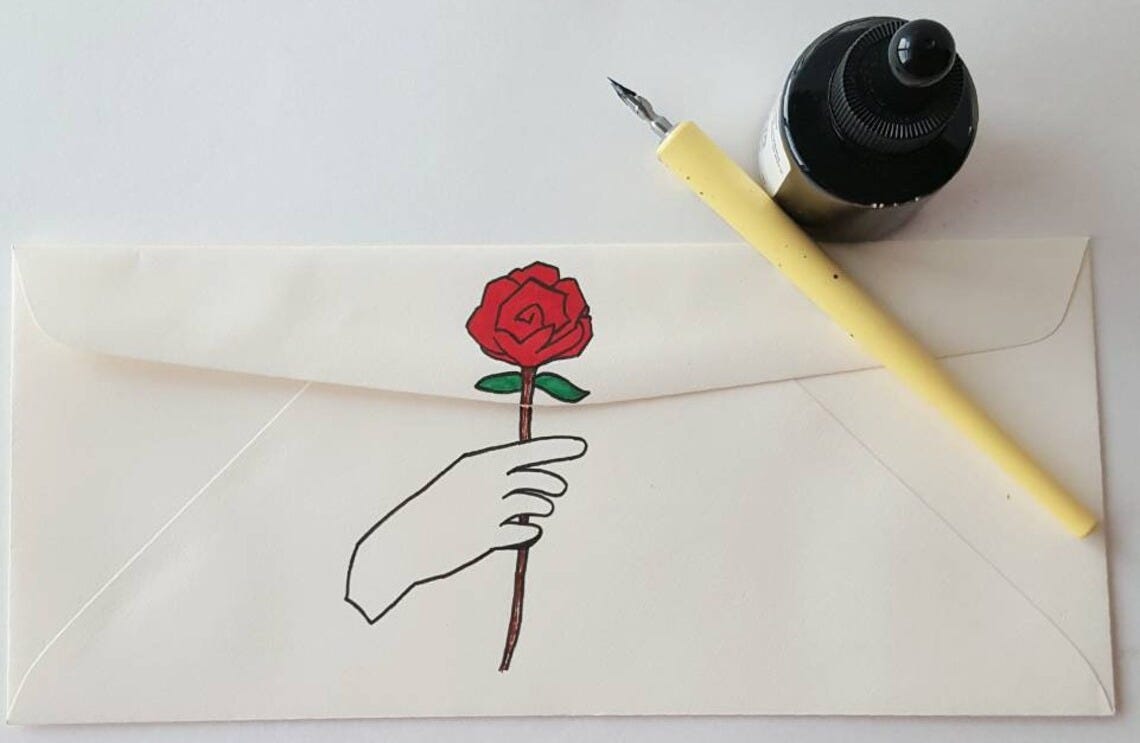 May include: A white envelope with a black outline of a hand holding a red rose with a green stem and leaves. A yellow calligraphy pen and a black ink bottle are on top of the envelope.