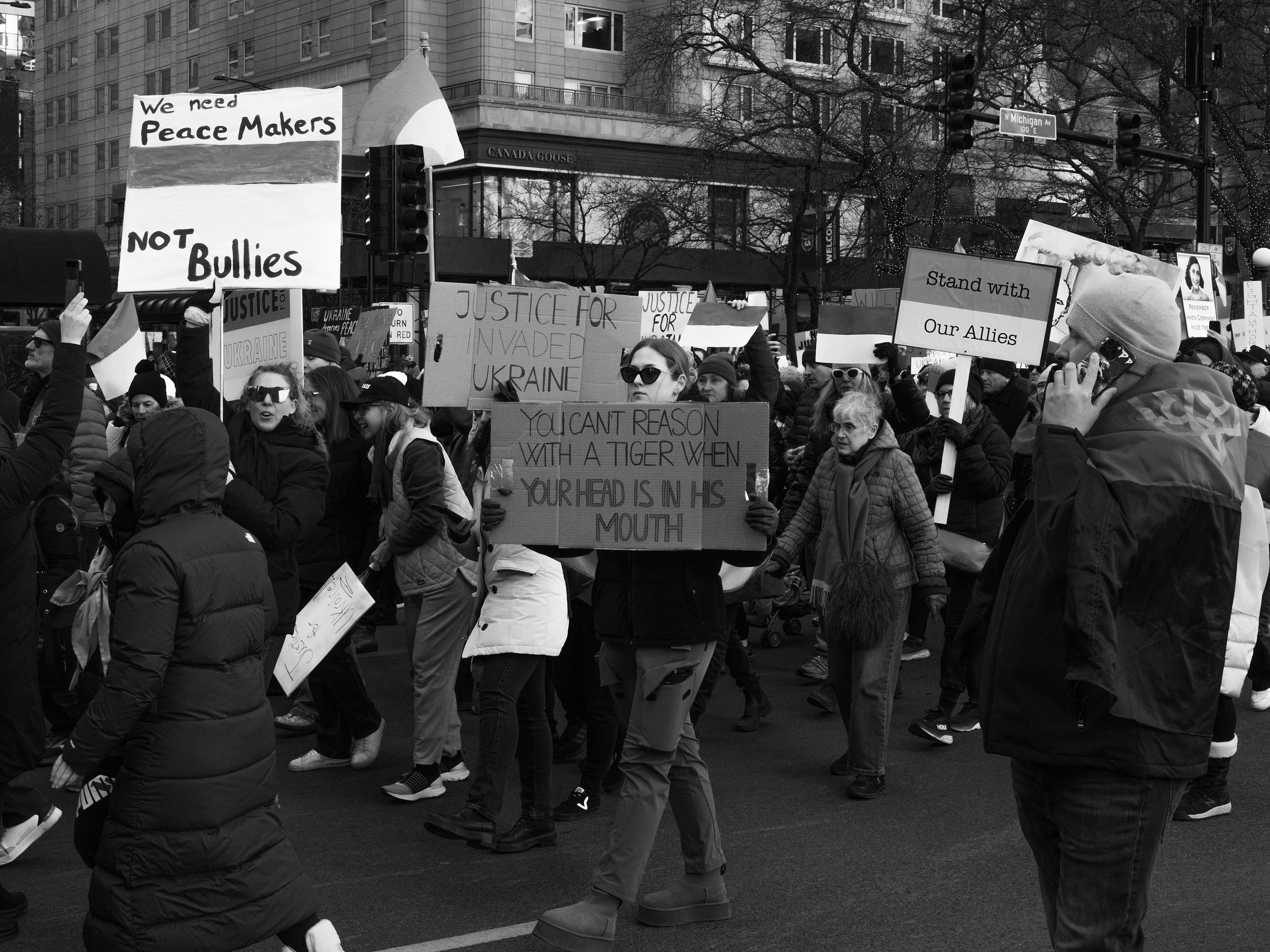 A black and white photograph captures a protest on a city street with demonstrators holding signs advocating for Ukraine. One prominent sign reads, "Justice for Invaded Ukraine – You can't reason with a tiger when your head is in his mouth." Another sign states, "We need Peace Makers, NOT Bullies." The crowd, dressed in winter clothing, marches with urgency, while a man wrapped in a flag talks on his phone. Urban buildings, traffic lights, and more protest signs fill the background, highlighting the energy and determination of the movement. A black and white photograph captures a protest on a city street with demonstrators holding signs advocating for Ukraine. One prominent sign reads, "Justice for Invaded Ukraine – You can't reason with a tiger when your head is in his mouth." Another sign states, "We need Peace Makers, NOT Bullies." The crowd, dressed in winter clothing, marches with urgency, while a man wrapped in a flag talks on his phone. Urban buildings, traffic lights, and more protest signs fill the background, highlighting the energy and determination of the movement.