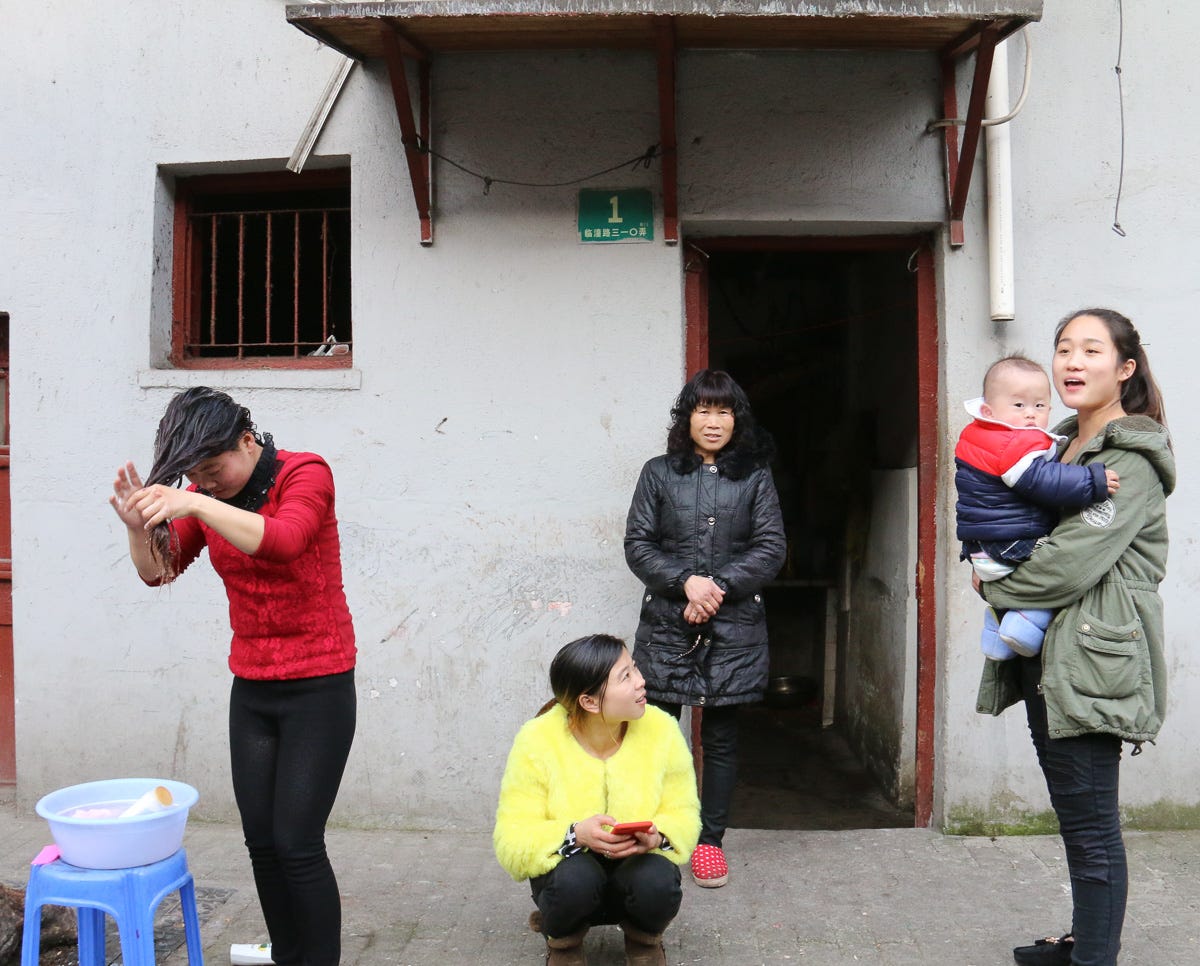 A woman washes her hair in a bucket while other women, including one holding a baby, gather in front of a home. A multi-generational street portrait of daily life.