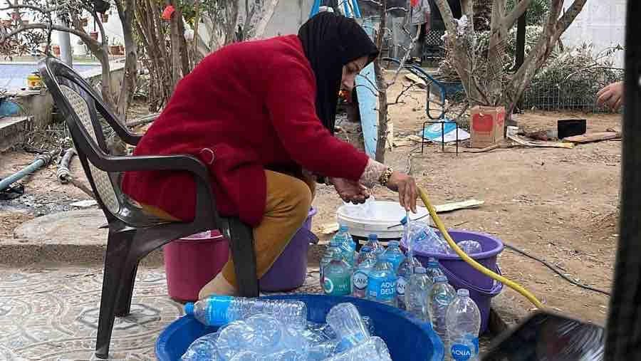 A woman wearing a red coat and black headscarf sits on a plastic chair outdoors, filling empty plastic water bottles using a hose. Several basins and containers filled with bottles are arranged on the ground around her in a sparse, dirt yard with trees and household items in the background.