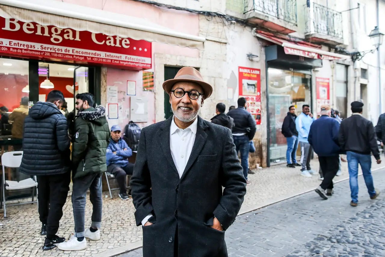 Un uomo sorridente con gli occhiali, un cappello marrone e un cappotto scuro sta in piedi in una strada acciottolata di Lisbona. Sullo sfondo si vede il "Bengal Cafe" con diverse persone davanti all'ingresso e lungo il marciapiede della via cittadina.