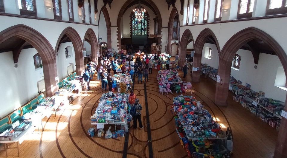church hall set up with rows of tables filled with children's items for sale. Crowd of people browsing