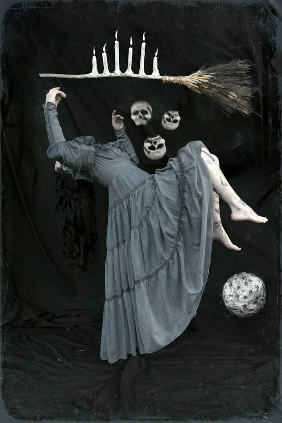 A surreal, dark-themed photograph featuring a figure in a flowing grey dress suspended in mid-air. The figure's head is replaced by four floating skull-like objects. Above, a broomstick with lit white candles hovers horizontally. A glowing orb sits on the ground below. The background is black, creating a stark contrast with the pale elements in the scene. A surreal, dark-themed photograph featuring a figure in a flowing grey dress suspended in mid-air. The figure's head is replaced by four floating skull-like objects. Above, a broomstick with lit white candles hovers horizontally. A glowing orb sits on the ground below. The background is black, creating a stark contrast with the pale elements in the scene.