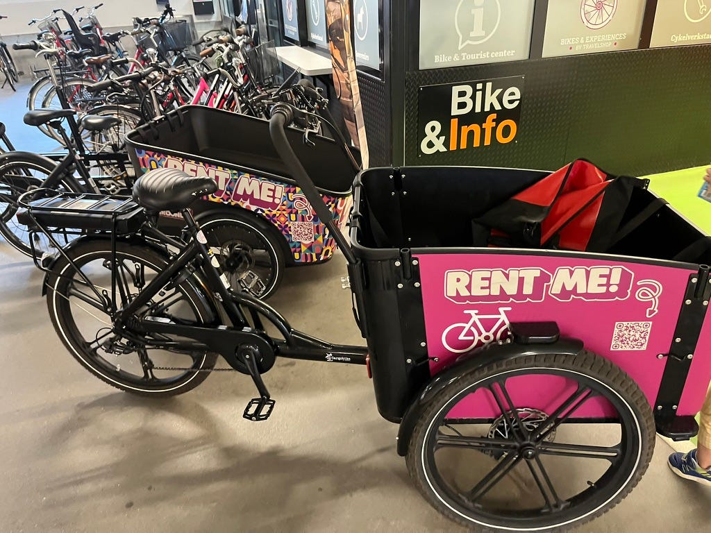 2 front-loader cargo bikes, each of which says "RENT ME!" on the colorful bucket. There are regular bikes behind them.