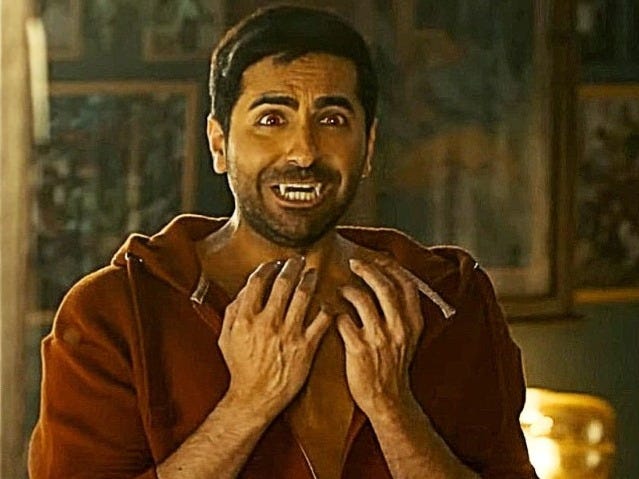 A still from Hindi film, ‘Thamma’. Alok (Ayushman Khurana) in a rust-colored hoodie is standing in a room, clawing his hands in front of his chest; his eyes are wide, brows raised, and mouth with fanged teeth open in a strained grimace. He is panicking. Warm light falls across his face, and the blurred background shows framed pictures on dark walls.