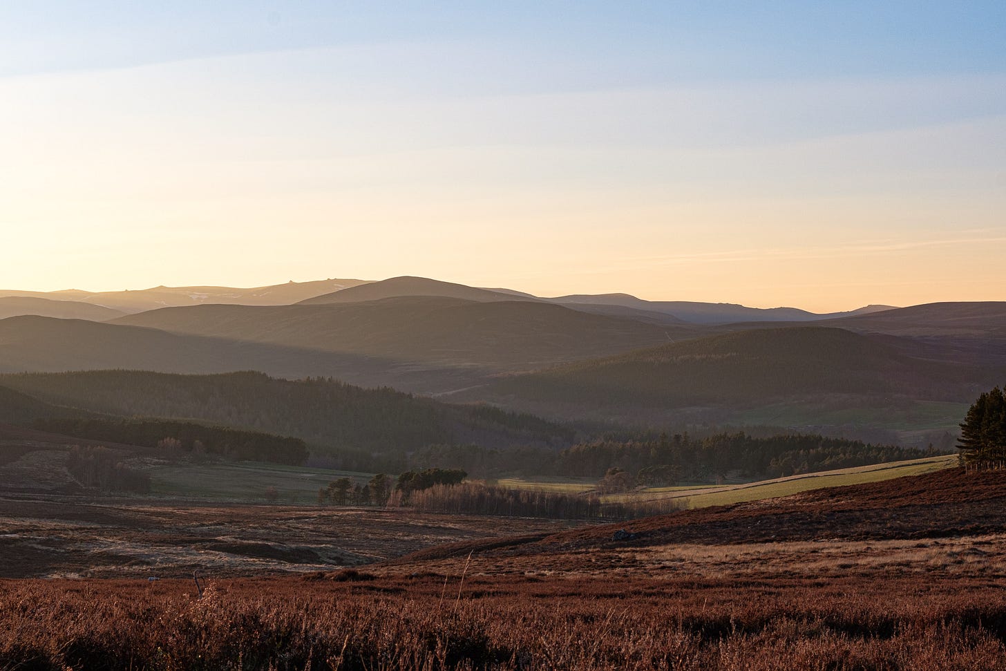 Rolling hills stretch across the Cairngorms National Park, their heather and forests bathed in golden light from the setting sun. Beams of sunlight break through the valleys, creating a dreamlike, nostalgic scene.