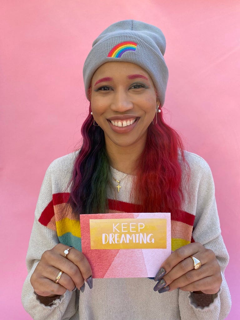 Kiana with rainbow hair wearing an American Eagle rainbow knit sweater holding up a handmade card that says Keep Dreaming