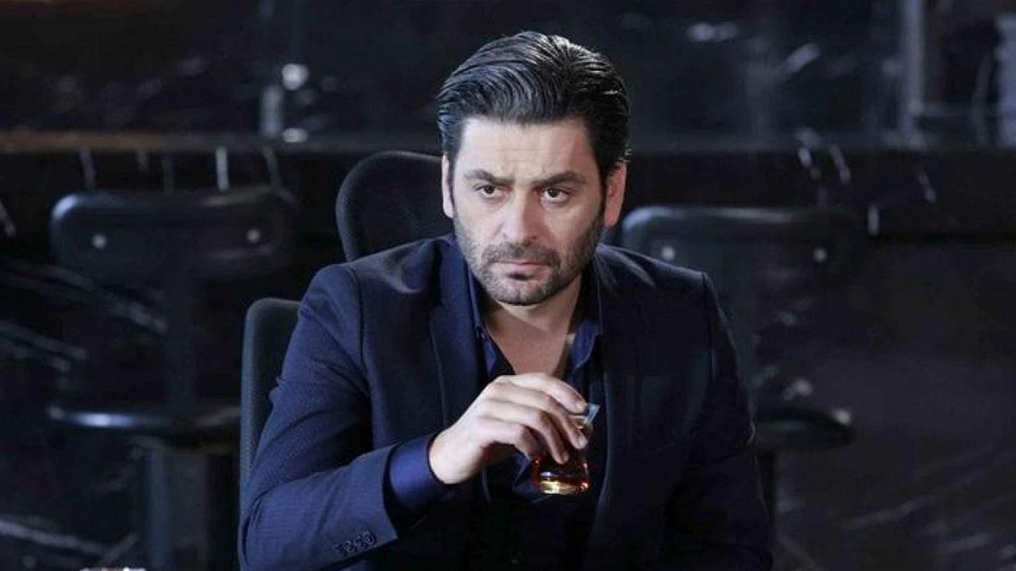 Eşkıya Dünyaya Hükümdar Olmaz dizisinde İlyas Çakırbeyli karakterini canlandıran Ozan Akbaba elinde çay bardağı ile masada otururken.