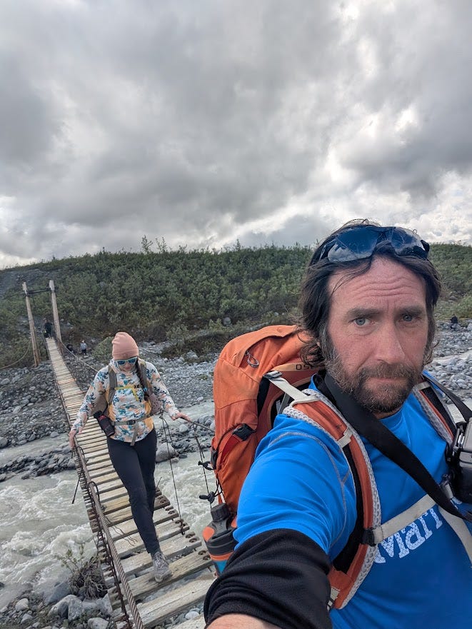 A selfie taken mid-crossing on a narrow wooden suspension bridge over a milky grey glacial river. A bearded man in a blue shirt with an orange pack and sunglasses pushed up on his head looks into the camera, tired-eyed. Behind him, a girl in a pale patterned hoody, beanie, and sunglasses steps carefully along the single-file planks, holding the cable rails on both sides. Heavy grey clouds hang over a brushy green hillside beyond.