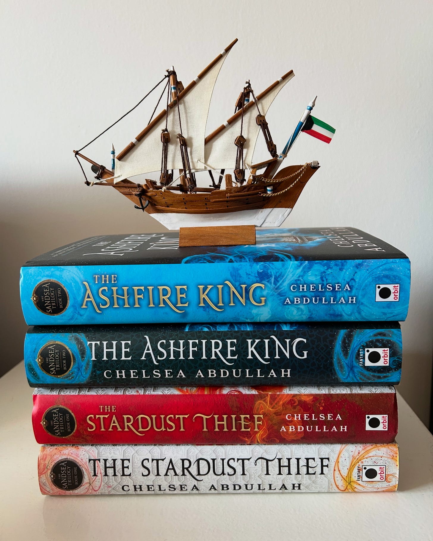 A model dhow with a Kuwaiti flag sits atop a stack of books: Two copies of THE ASHFIRE KING with the US and UK spines and two copies of THE STARDUST THIEF with a red and white spine.