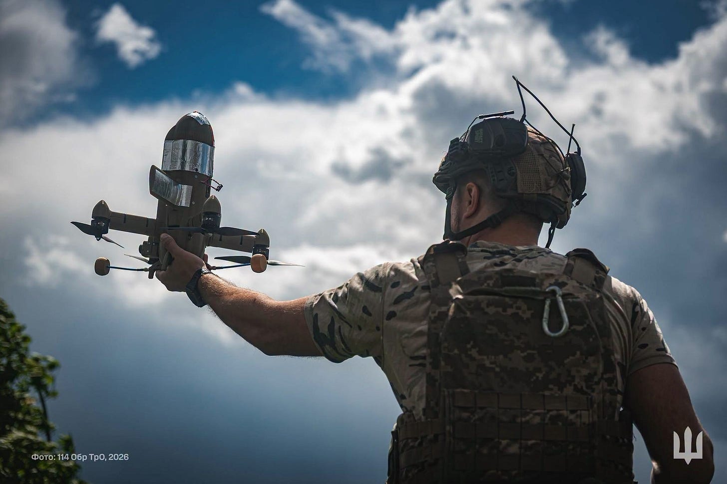 A soldier in camouflage and helmet holds up a small fixed-wing interceptor drone against a cloudy sky, seen from behind