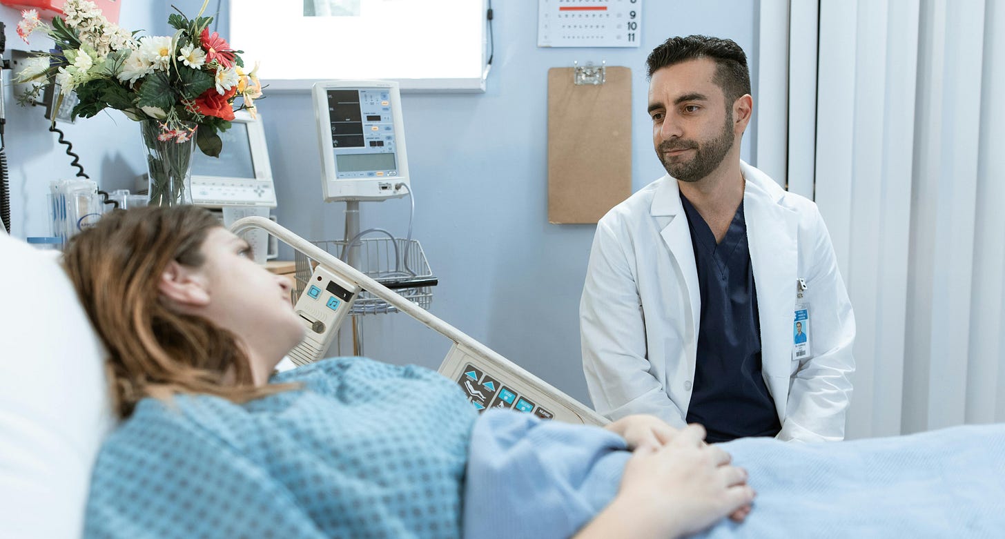 A patient, lying in a hospital bed in the foreground, talks to a doctor, sitting, in the background. They are both in a hospital room.